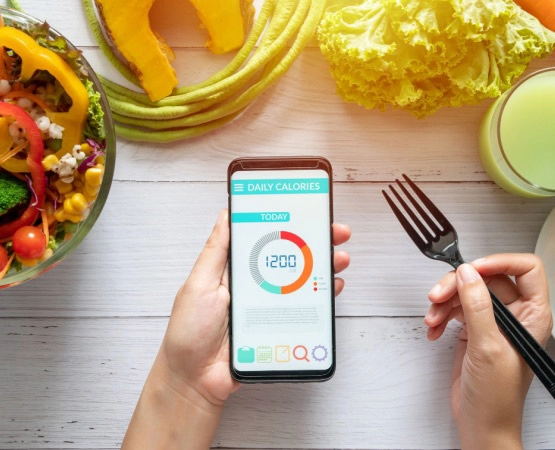 Woman using calorie counter application on her smartphone at dining table with salad, fruit juice, b